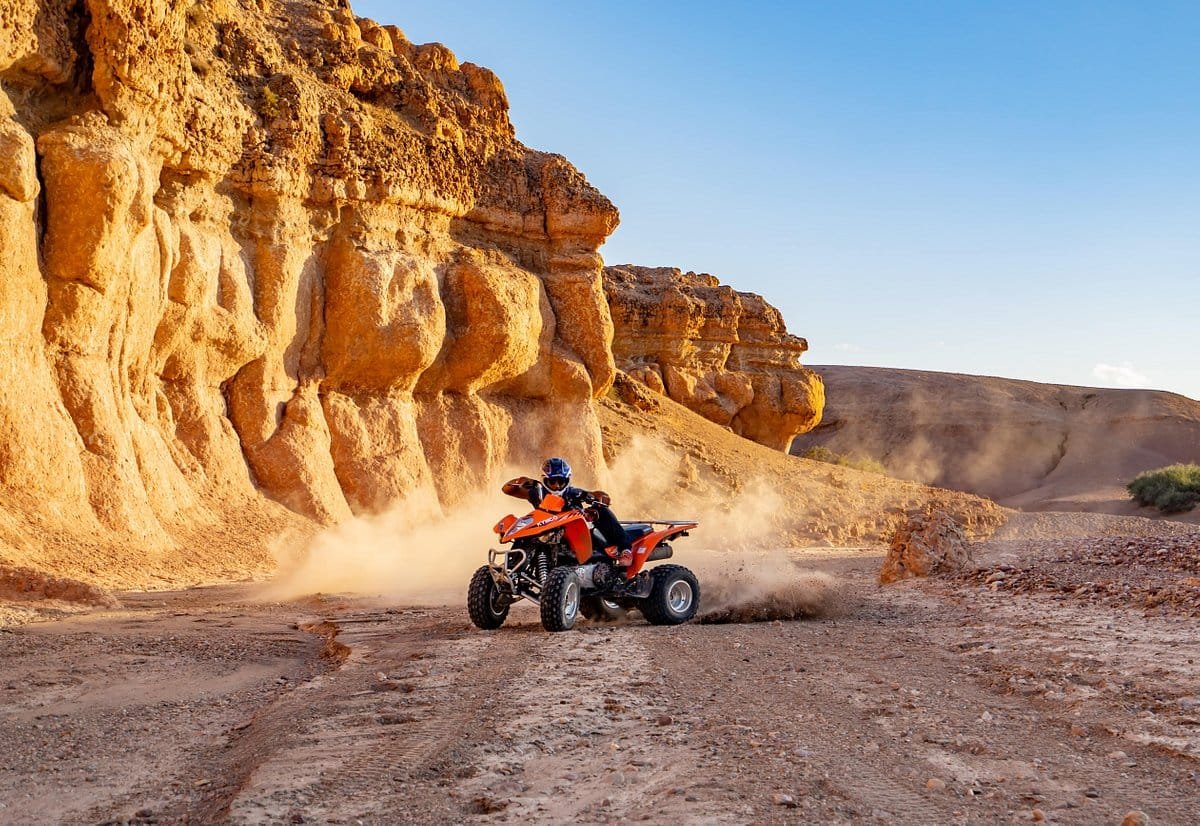 riding quad bikes through the Agafay Desert near Marrakech, kicking up dust as they navigate the sandy terrain. The riders are wearing protective gear and helmets, and the desert landscape stretches out in front of them with a hazy mountain range visible in the distance.