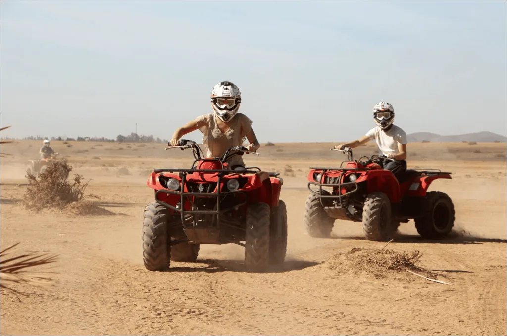 quad bikes in the Agafay Desert near Marrakech. The sun is setting in the background, casting a warm orange glow across the sand dunes. The riders are wearing helmets and protective gear, and appear to be having a thrilling adventure