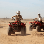 quad bikes in the Agafay Desert near Marrakech. The sun is setting in the background, casting a warm orange glow across the sand dunes. The riders are wearing helmets and protective gear, and appear to be having a thrilling adventure