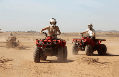 quad bikes in the Agafay Desert near Marrakech. The sun is setting in the background, casting a warm orange glow across the sand dunes. The riders are wearing helmets and protective gear, and appear to be having a thrilling adventure