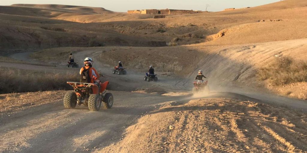 People riding quad bikes in the Agafay Desert Marrakech, with rocky terrain and hills in the background. The sun is shining bright and the riders look excited and adventurous.