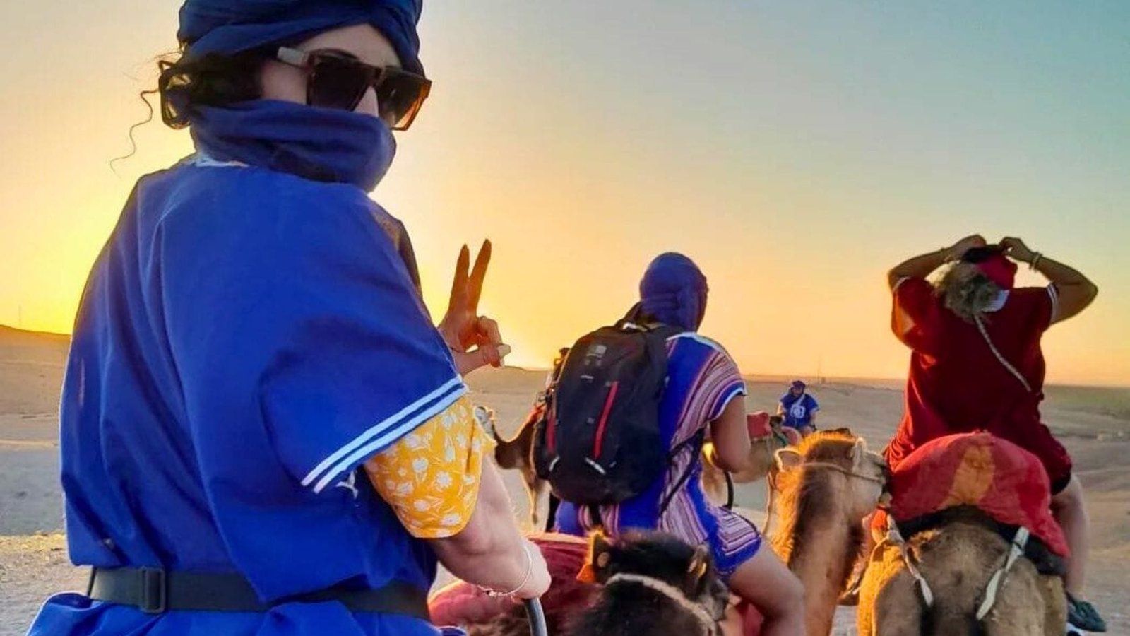 Group of travelers riding camels in the Agafay Desert at sunset, wearing traditional blue and red outfits.