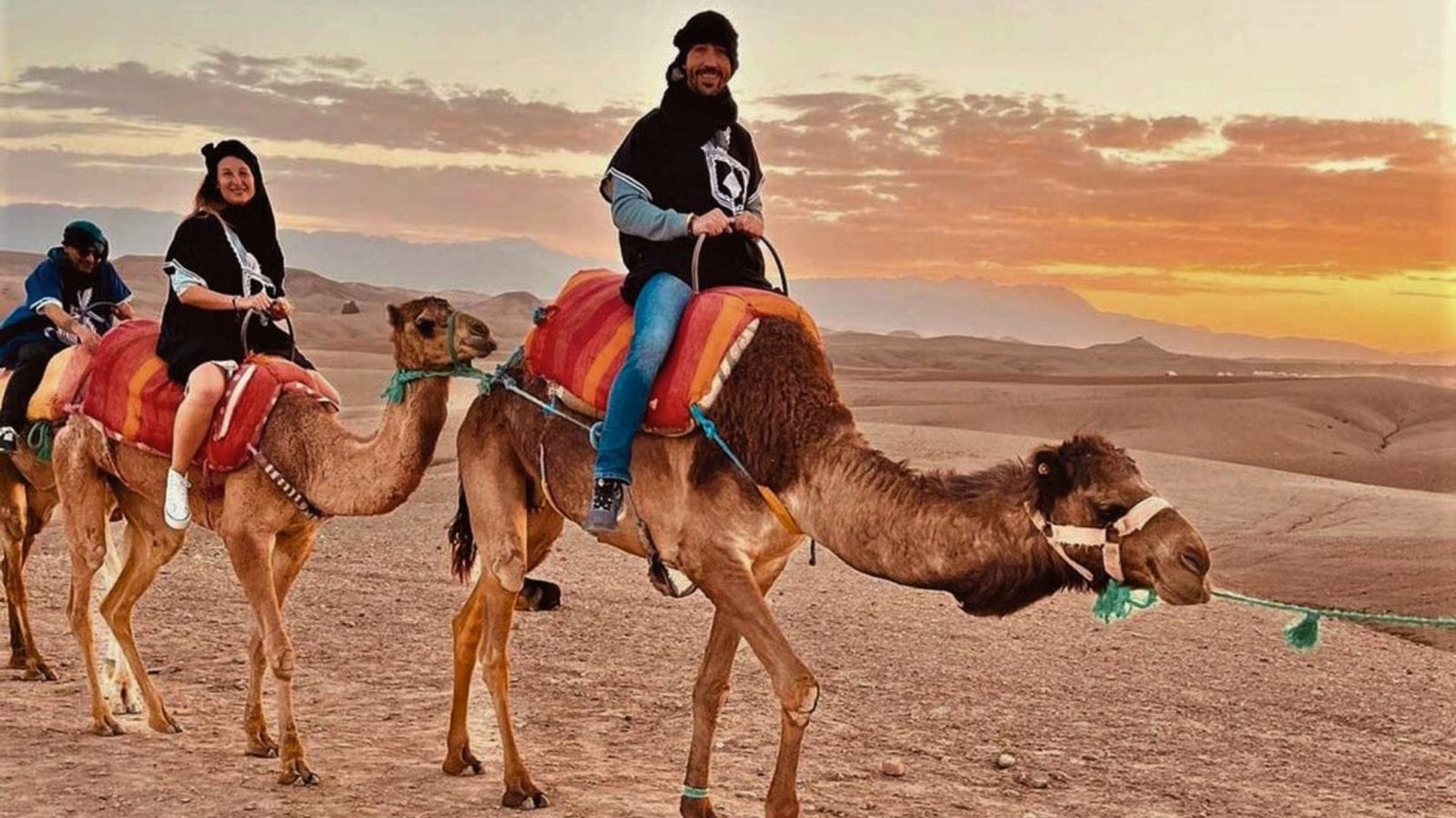 Couple riding camels at sunset in the Agafay Desert with glowing orange skies.